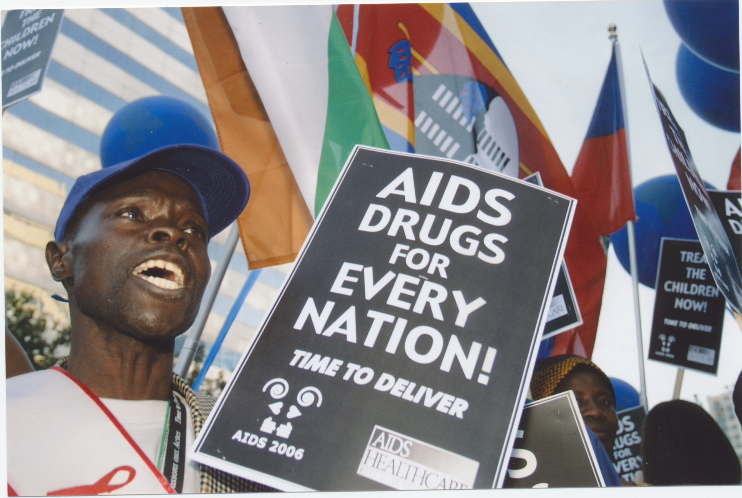 A person holding a sign that reads “AIDS Drugs for Every Nation! Time to Deliver” amidst flags and banners, advocating for global access to AIDS medication.