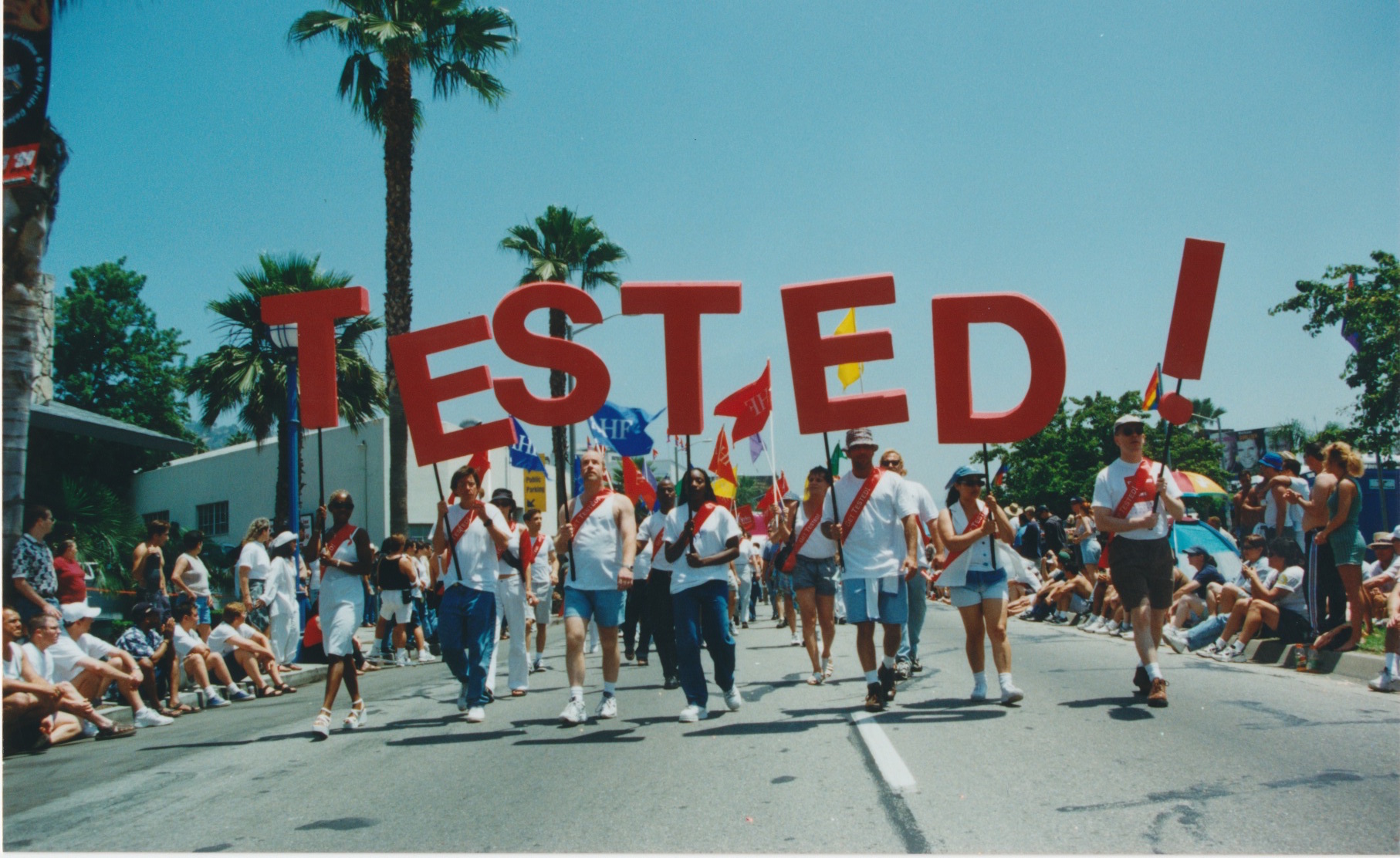 A group of people marching in a parade holding large red letters spelling “TESTED!”